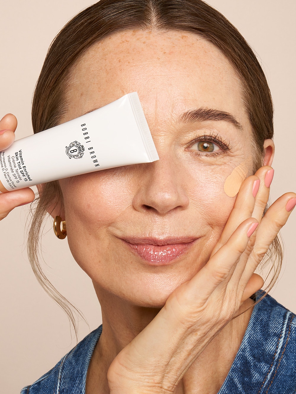 Mature model playfully covering one eye with new Vitamin Enriched Skin Tint, in front of a bright yellow background